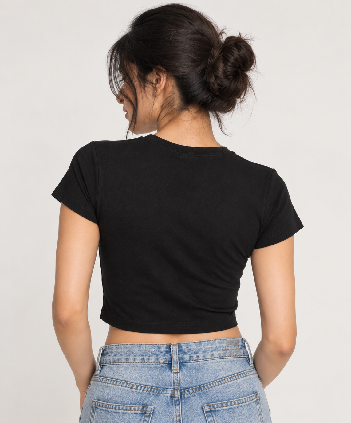 A woman shown from the back wearing a fitted black short-sleeve crop top and light blue jeans against a plain light gray studio background.